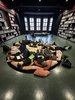 Eine Gruppe von jungen Leuten liegt auf dem Boden inmitten von unterschiedlich großen und bunten Kissen. Der Raum ist seitlich gefüllt mit Bücherregalen. Am Ende des Raumes sieht man die Sprossenfenster des Raumes.