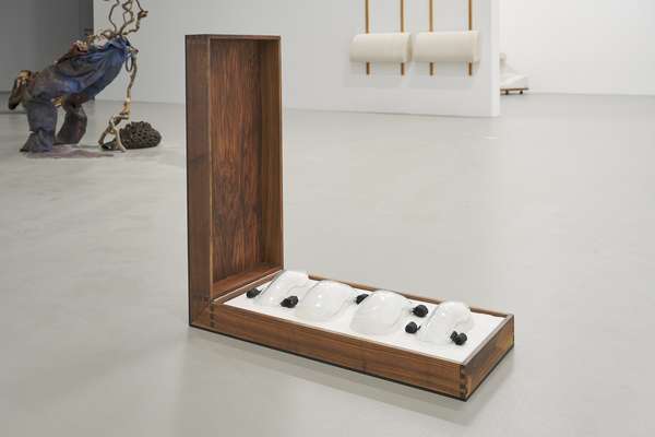 Im Fokus des Bildes steht eine geöffnete Holzbox mit vier weißen Glasobjekten und zwischen ihnen mehrere kleine schwarze Objekte. Im Hintergrund sind weitere Objekte im Raum zu sehen. Die Kiste steht auf dem Boden.