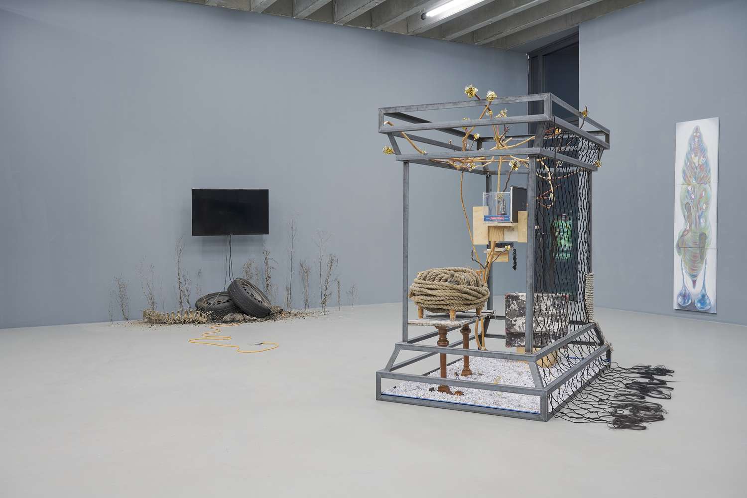 Im Vordergrund des Ausstellungsraumes steht eine Übermenschen große Skulptur aus unterschiedlichsten Materialien, die in einem Metallrahmen gefasst ist. Im Raum dahinter hängt ein Bildschirm an der Wand, darunter Autoreifen und Andeutungen von vegetabilen Strukturen.