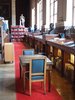 Es ist eine Bibliothek zu sheen: Bücher in Regalen, ein roter Teppich führt vom Bildvordergrund in den -hintergrund. Darauf steht im Vordergrund ein Lesetisch und zwei lederbepolsterte Stühle.