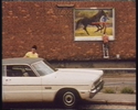 Vor einer großen Ziegelwand mit einer Plakatfläche steht ein altes, weißes, amerikanisches Auto. Vor der Plakatwand steht eine Leiter, eine Person steht darauf. Hinter dem Auto, anscheinend die Fahrertür öffnend, steht eine Frau im gelben TShirt.