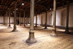 Das Bild zeigt einen großen Raum mit einem Holzdielenboden. Der Raum wird durch eine Vielzahl an schmalen Säulen aus Metall durchbrochen. Es herrscht durch die Beleuchtung eine schummerige Atmosphäre.