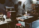 Eine dunkle Werkstatt/Atelier wird gezeigt mit vielen Dingen, Tischen, Kleinteilen. Ein altes Faxgerät steht im Vordergrund, daneben ein alter Aktenschrank mit einem alten, recht großen Vogelhaus. An der Wand hängen weitere Gegenstände, wie ein Hochkant-Porträt eines Mannes im Trenchcoat neben einem Hirschgeweih.