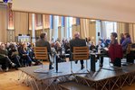 Man sieht die Podiumsteilnehmer und -teilnehmerinnen von hinten. Der Blick wird frei auf das Publikum in der Aula.