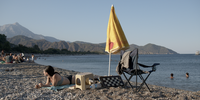 Eine Frau liegt bäuchlings an einem Kiesstrand. Rechts neben ihr stehen ein Campingstuhl und ein gelber Sonnenschirm, der nicht aufgespannt ist. Im Hintergrund ist ein See oder das Meer zu erkennen.