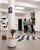 A group of students is standing in an exhibition room looking at art. A black and white chequered blanket lies on the floor, next to it is a picnic basket. Black, star-shaped structures are stuck to the white walls, and a wooden desk stands in front of them. Pieces of wood have been mounted around a column in the foreground of the picture so that they hold a bouquet of flowers. 