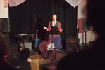 Eine Frau steht auf einer Bühne mit diversen Musikinstrumenten. Vor ihr steht ein Mikro, in das sie hinein singt.