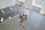 Aus einer erhöhten Perspektive sieht man zwei Besucher*innen mit durchsichtigen Regenschirmen über ein Stahlgitter laufen. Es nieselt im Raum.