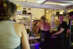 Menschen stehen in einer Würstchenbude. Man sieht eine Frau in Rückenansicht am Tresen, die sich mit einem Mann unterhält.