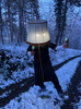 Ein Mann steht mit einem Lampenschirm auf dem Kopf in einem winterlichen Wald, samt Schnee auf dem Weg.