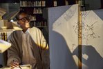 Auf der rechten Seite ist eine Frau zu sehen mit großer schwarzer Brille, die von dem Licht von der vor ihr stehenden Overhead-Projektoren indirekt angestrahlt wird. Auf der rechten Seite des Bildes werden die projizierten Zeichnung auf einer weißen Wand dargestellt.
