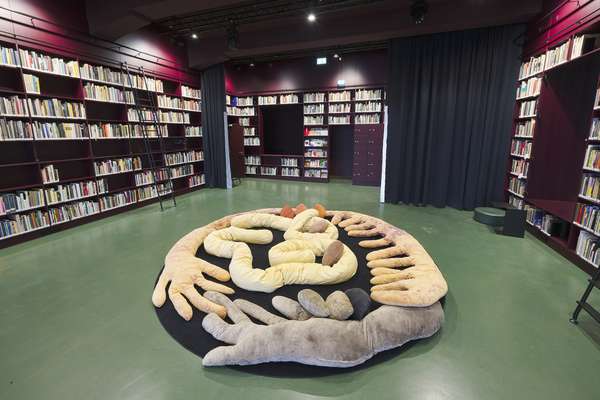 Rechts und links wird der Raum durch Bücherregale gerahmt. In der Mitte sind unterschiedlich große und farbige Kissen auf einem runden Teppich ausgelegt.