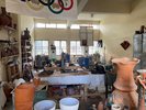 Der Blick in einer Keramik-Werkstatt: um einen Tisch in der Mitte des Bildes stehen diverse Regale und andere Abstellmöglichkeiten mit unterschiedlich großen und gestalteten Keramik-Stücken.