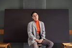Eine Frau mit einem Kurzhaarschnitt ist in einen grauen, karierten Blazer mit einem orangenen Rolli gekleidet und sitzt vor einer schwarzen Wand. Sie trägt außerdem eine Brille mit einem hellen Metallgestell und hat dabei ihre Hände im Schoß leicht gefaltet. Ihr Kopf ist geneigt und schaut direkt in die Kamera.