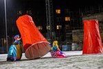 Two people in outlandish costumes move oversized red plastic cups on a snow-covered sports field