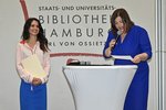 Links steht eine lächelnde Frau in blauen Rock, rosaner Bluse und langen braunen Haaren. Sie hält eine beige Papiermappe in der Hand. Rechts neben ihr steht eine weitere Frau in einem blauen Kleid, die ein Mikrophon zum Mund hält und von einem Blatt etwas abliest.