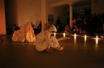 In Kerzenschein sitzt eine junge Frau in weißer Kleidung auf dem Boden in einem dunklen Raum. Im Hintergrund sind viele Besucher*innen zu sehen, die ihre Performance ansehen.