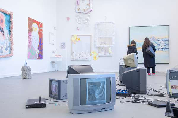 In the middle of the room, several old television sets are connected by cables. Large, colourful pictures hang on the walls. On the right of the picture, two women are standing in front of an abstract mountain landscape in pastel shades.