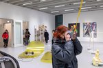 In the foreground, a red-haired woman looks at a work of art through VR glasses hanging from the ceiling. In the background, several works are displayed on the floor and walls of the gallery in the new studio building. 