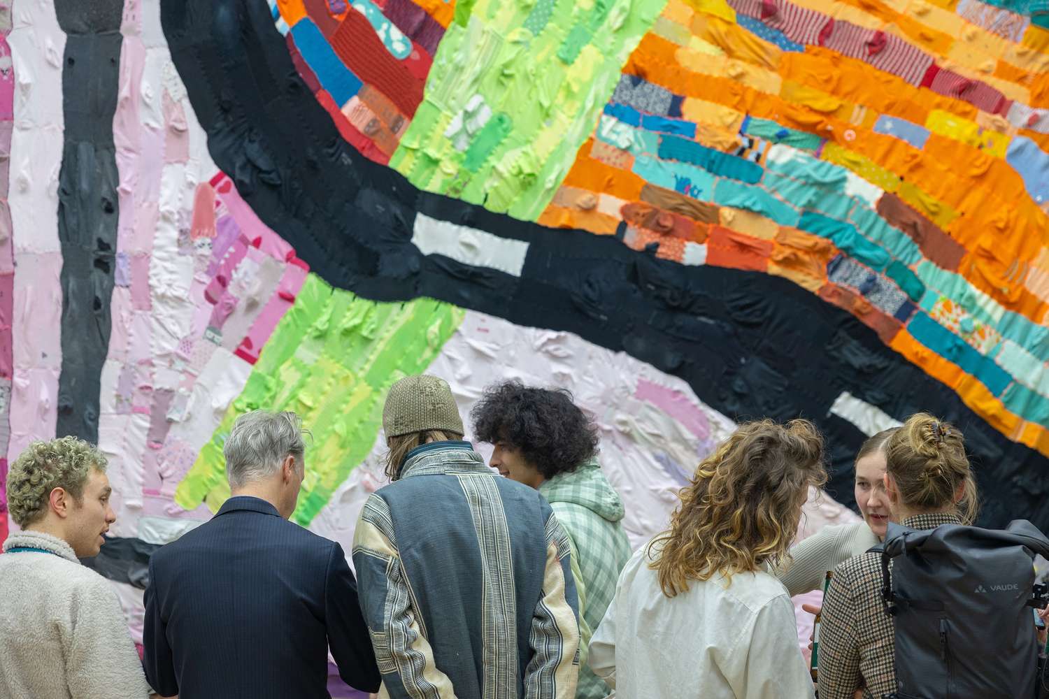 Sieben Personen stehen vor einer bunten Wand aus unterschiedlich farbigen Stoffstreifen.