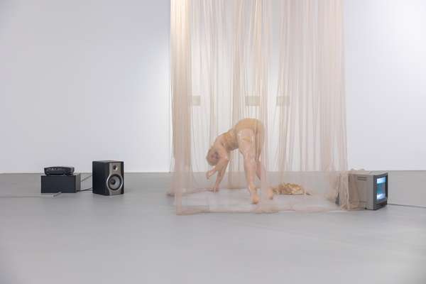 In einem weißen Raum stehen Kleinelektronikgeräte auf dem Boden. Mittig steht eine weibliche Person hinter einem beigen Schleier auf ihren Fußspitzen, sie ist nach vorn gebeugt und stützt sich mit einer Hand auf dem Boden ab.