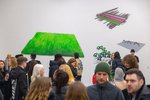 Viele Menschen befinden sich in einem Ausstellungsraum und reden miteinander. An der Wand sind Kunstwerke in unterschiedlichen Formaten und Farben zu sehen. Eines erscheint wie eine neon-grüne Wiese.