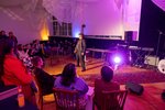 Ein in farbiges Licht getauchter Raum ist mit einem Publikum gefüllt, dass sich eine Performance ansieht: ein Mann steht gestikulierend vor dem Publikum.