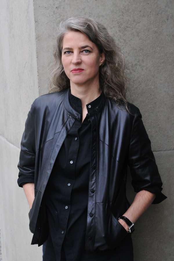 Die Porträtaufnahme einer Frau mit dunklen, lockigen Haare, die vereinzelt schon ergraut sind. Sie trägt eine schwarze Jacke und roten Lippenstift. Sie hat ihre Hände in den Jackentaschen.