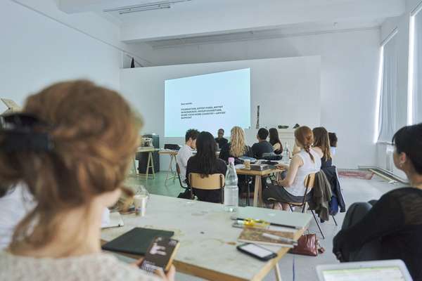 Ein Raum voller junger Studierender wird gezeigt. Vor der Projektion an der hinteren Wand ist eine dunkelhäutige Frau mit schwarzer Brille und ebenso schwarzer Bluse zu sehen. Sie ist die Lehrbeauftragte des Seminars und schaut ihre Studierenden mit wachen freundlichen Augen an.