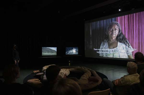In einem dunklen Raum wird ein Film an die rechte Wand projiziert.. Eine indigene Frau schaut in die Kamera. Im Vordergrund liegen Besuchende auf Kissen. Im Dunkeln sind zwei weitere Bildschirme zu erkennen.