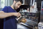 Ein Mann mit Dreitagebart, schwarzen Haaren und einem blauen T-Shirt bereitet einen Kaffee an einer Gastromaschine zu.