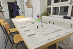 Ein selbstgemaltes Spielbrett, Stifte, Papierblöcke liegen auf einem Tisch. Daran gestellt wurden mehrere Stühle. Es ist hell in dem Raum. Der Tisch steht an einem Fenster mit einer Heizung.
