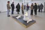 Im Vordergrund ist ein skulpturale Installation zu sehen, die hauptsächlich aus der Gipsabformung einer weiblichen Gestalt besteht. Besucher*innen der Ausstellung stehen um die Installation herum und betrachten diese. Andere Besucher*innen bewegen sich im hellen  Raum.