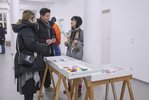 Drei Menschen unterhalten sich in einem hellen Ausstellungsraum. Vor ihnen in Schubladen aufgebockt bunt gefaltete Umschläge.