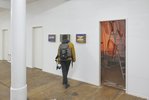 Eine Person steh vor drei kleinformatigen im Querformat gehalten nBildern, die an einer weißen Wand hängen. Rechts von der Person öffnet sich eine Tür in einen Raum, der durch eine durchsichtige Plastikwand vom Hauptraum getrennt wird. Dahinter kann man eine große Klappleiter erkennen. Der Raum ist in oranges Licht getaucht.