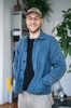 Ein junger Mann mit Brille schaut in die Kamera und lächelt mit leicht geöffnetem Mund. Er trägt eine blaue Jacke, darunter ein schwarzes Oberteil. Seine Hände hat er in die Jackentaschen gesteckt.
