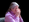 Ein Filmstill wo eine Frau mit grauem längerem Haar sich die Augen verdeckt, der Hintergrund ist Schwarz, man kann noch ihren Sessel erkennen, sie trägt einen rosafarbenen Pullover.