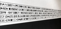 Auf weißer Wand sehen wir überlappende Buchstaben in schwarzer Schrift, auf Linie geschrieben. Der Boden ist schwarz.