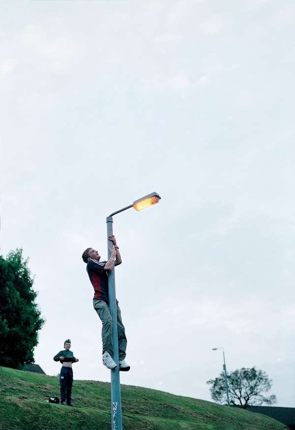 Ein junger Mann ist an einer Laterne hinauf geklettert. Der obere Bildrand ist mit weiß-blauen Himmel gefüllt.