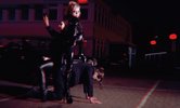 Das Filmstill zeigt eine Aufnahme in der Nacht auf einem Parkplatz, drei Personen in schwarzer Kleidung, leicht verschwommen und nicht klar zu sehen wie sie beieinander stehen
