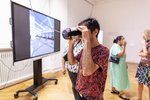 Ein Besucher im Vordergrund hat sich eine VR-Brille aufgesetzt. Im Hintergrund ist ein großer Bildschirm zu sehen, der darstellt, was der Besucher in diesem Augenblick sieht.