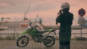 A motorcyclist in full gear has parked his bike by a river in front of an industrial harbour backdrop and is taking pictures of it.