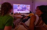 Zwei Besucherinnen liegen auf dem Boden zwischen Kissen. Das Foto wurde mit einem Blick zwischen die Schultern der beiden Schultern aufgenommen. Der Blick geht auf eine Videoleinwand.