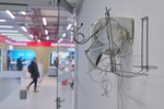 An einer weißen Wand hängt eine fragil aussehende Skulptur aus Metall und Glasgewebe, im Hintergrund sieht man verschwommen eine offene Glastür und Menschen in einem Technikgeschäft