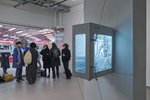 An einem weißen Pfeiler hängt ein silberfarbenes Computergehäuse, der Korpus ist beleuchtet und seitlich mit einer grauen Collage bedeckt. Im Hintergrund stehen mehrere junge Ausstellungsbesucher*innen