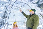 Ein Mann und ein kleines Mädchen stehen im Inneren eines Folientunnels und haben gelbe Brillen auf. Sie schauen nach oben und verfolgen mit ihren Blicken Federn, die durch den Tunnel wehen.