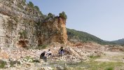 Ein schroffer Felsen erhebt sich über die linke Bildhälfte. Auf Bruchsteinen sitzen im Vordergrund drei Menschen. Im rechten hinteren Bildfeld erstrecken sich bewaldete Hügel. Es sieht trocken und warm aus.