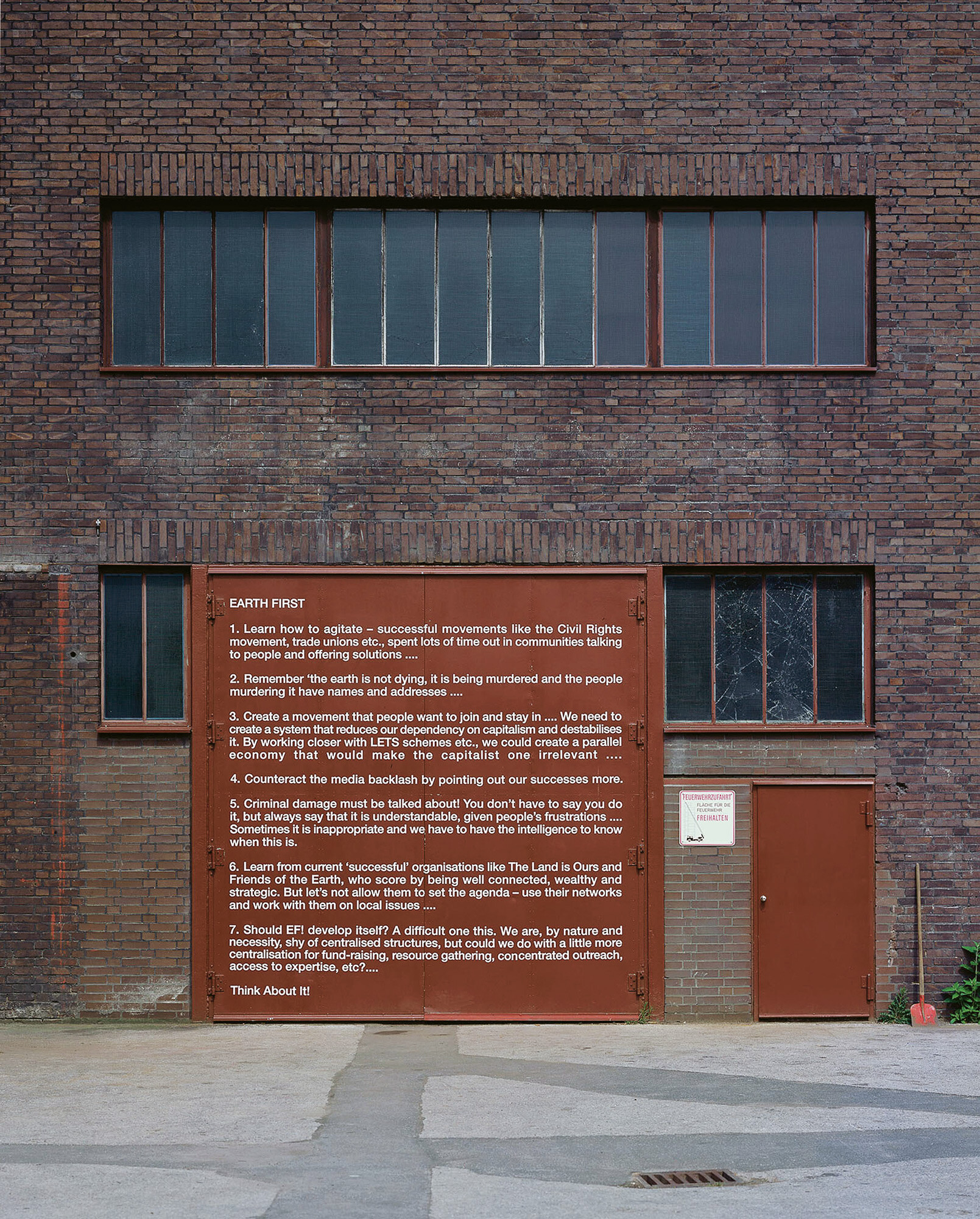 Brick facade of an industrial building with a long, narrow window on the upper floor. On the ground level there is a large red information sign with white text and a red metal door next to it. The ground in front is concrete.