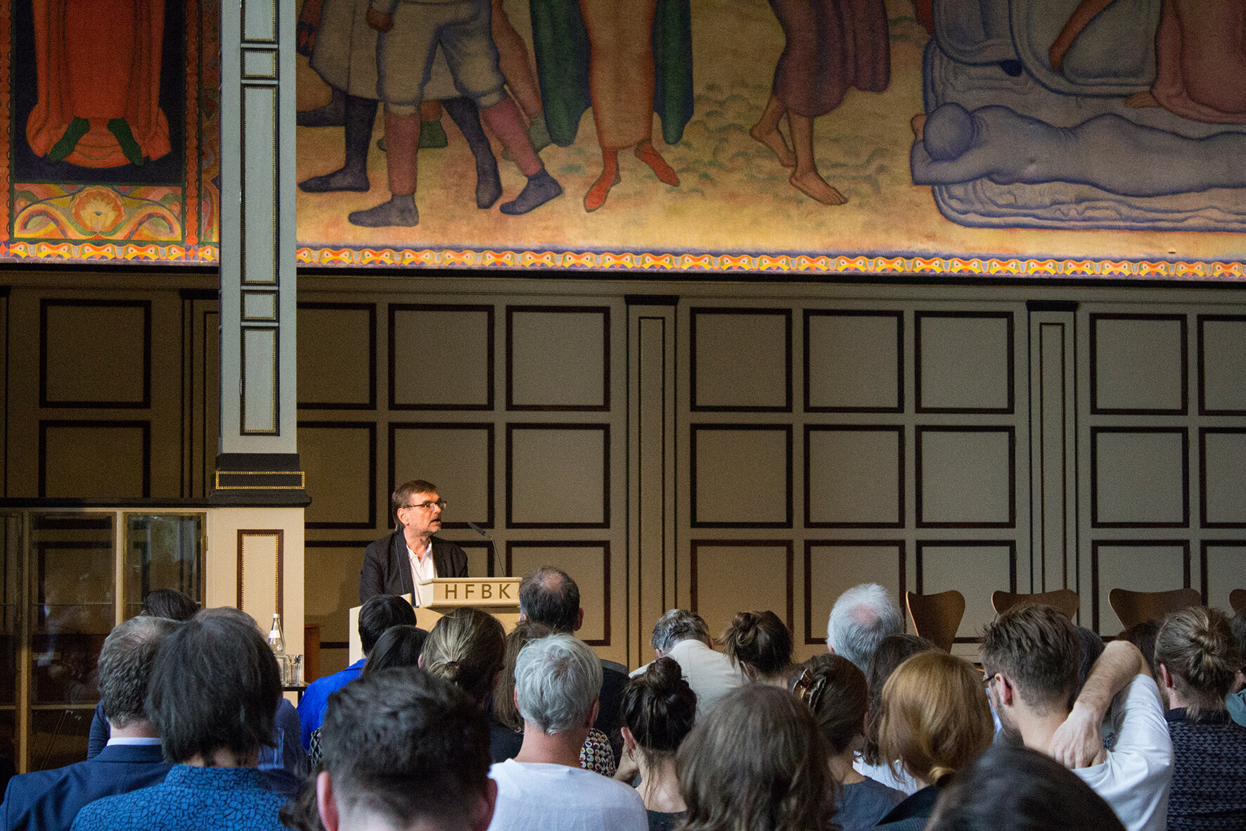 Lecture in the HFBK auditorium: A person speaks at a lectern in front of a seated audience. A large mural is visible above the scene.