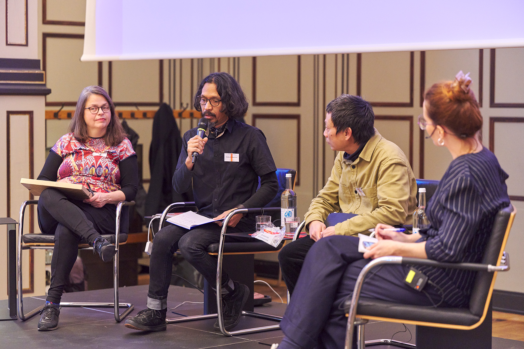 Podiumsgespräch in der Aula der HFBK Hamburg: Vier Personen sitzen auf der Bühne; eine Person spricht in ein Mikrofon, während die anderen zuhören und Notizen halten.